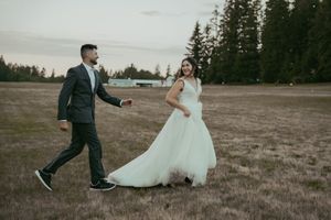 A bride in a white gown and groom in a dark suit walk hand in hand across a field at dusk.