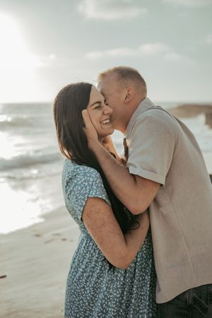 A couple shares a tender moment on a beach, man kissing woman's cheek while ocean waves crash behind them.