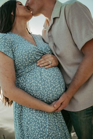 Pregnant couple sharing a kiss while cradling baby bump, woman in blue floral dress with engagement ring visible.