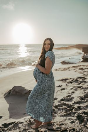 Smiling pregnant woman in floral blue dress cradling baby bump on sandy beach at golden sunset.