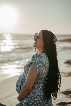Pregnant woman in floral dress holding belly on sunny beach at sunset.