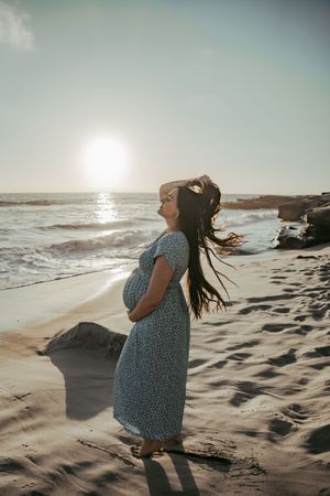 Pregnant woman in floral dress holding belly on sandy beach at sunset with ocean waves behind her.