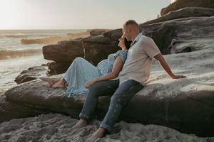 A pregnant couple sits intimately on rocky beach boulders at sunset, sharing a quiet romantic moment.