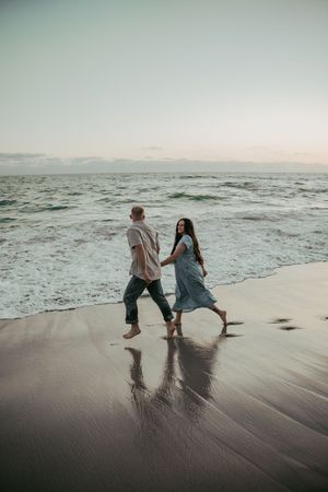 Couple holding hands walking barefoot on a wet sandy beach at sunset with ocean waves in the background.