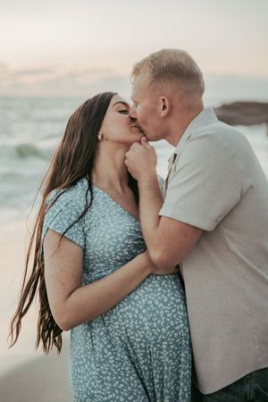 A couple shares a romantic kiss on the beach at sunset, woman in floral blue dress with long dark hair.