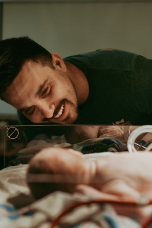 Happy father smiling at his newborn baby through the glass of a hospital bassinet.