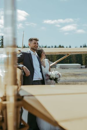 A bride and groom stand together near a small plane on a sunny day, with the bride holding a white floral bouquet.