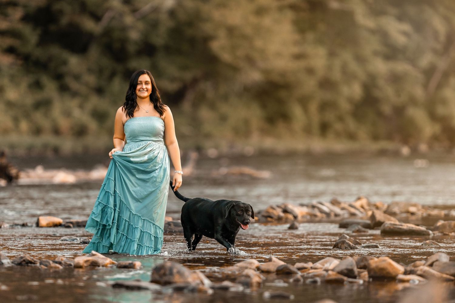 Senior girl in turquoise dress poses by rocky riverbank with black dog in scenic outdoor setting.