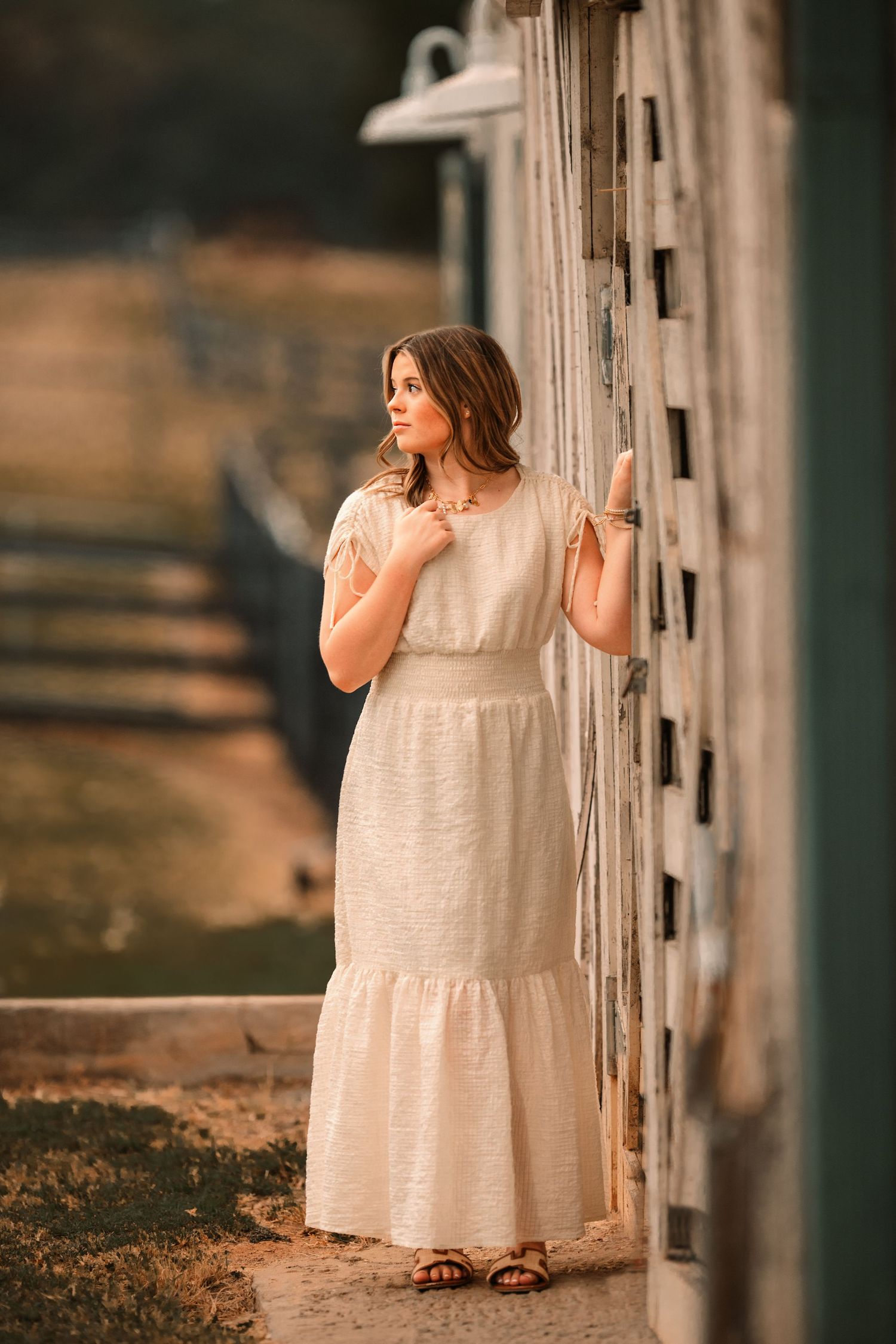 A senior in a white dress stands beside a wooden fence at sunset creating a dreamy golden hour portrait.