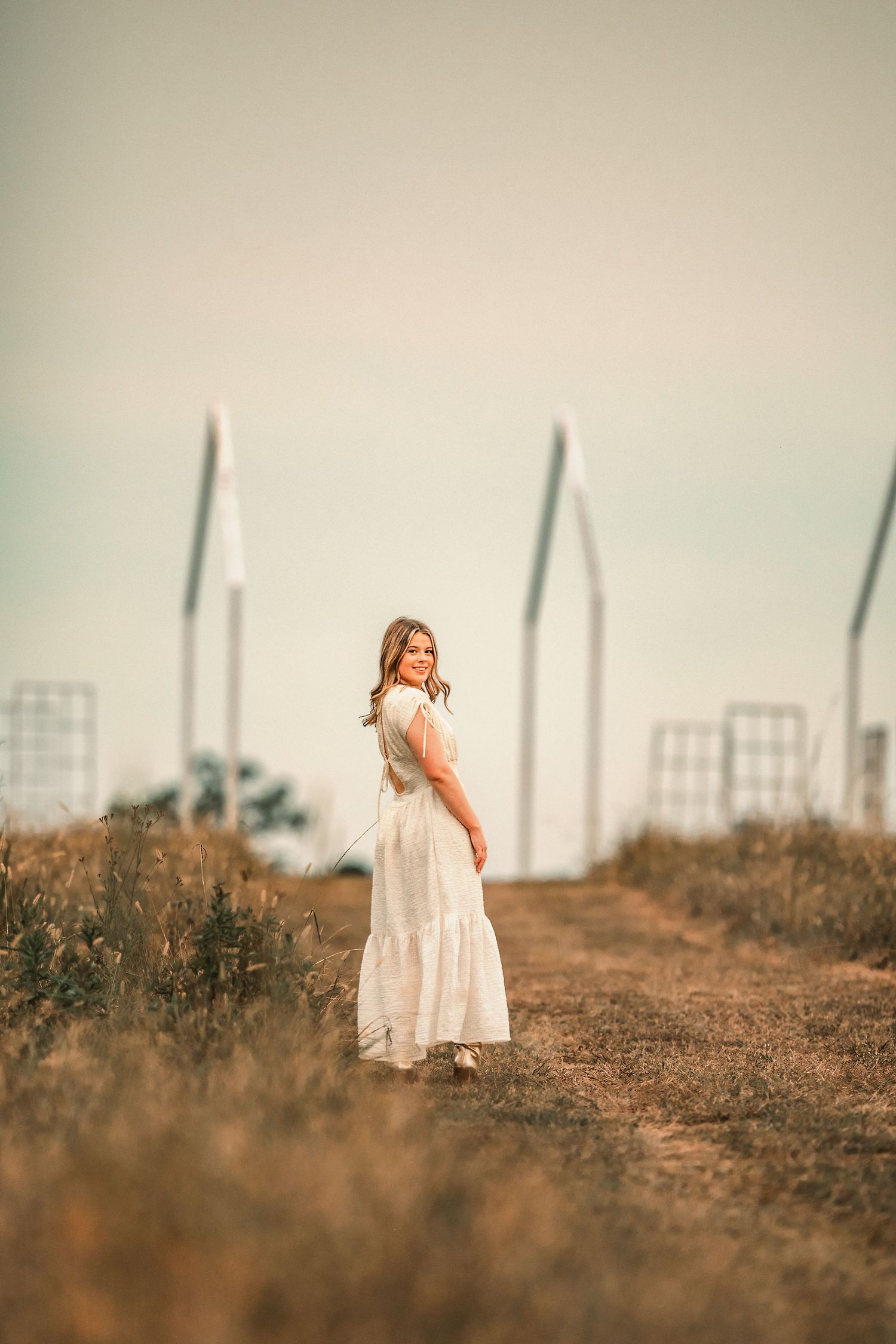 A high school senior in white dress stands in field during golden hour with dreamy, vintage effect.