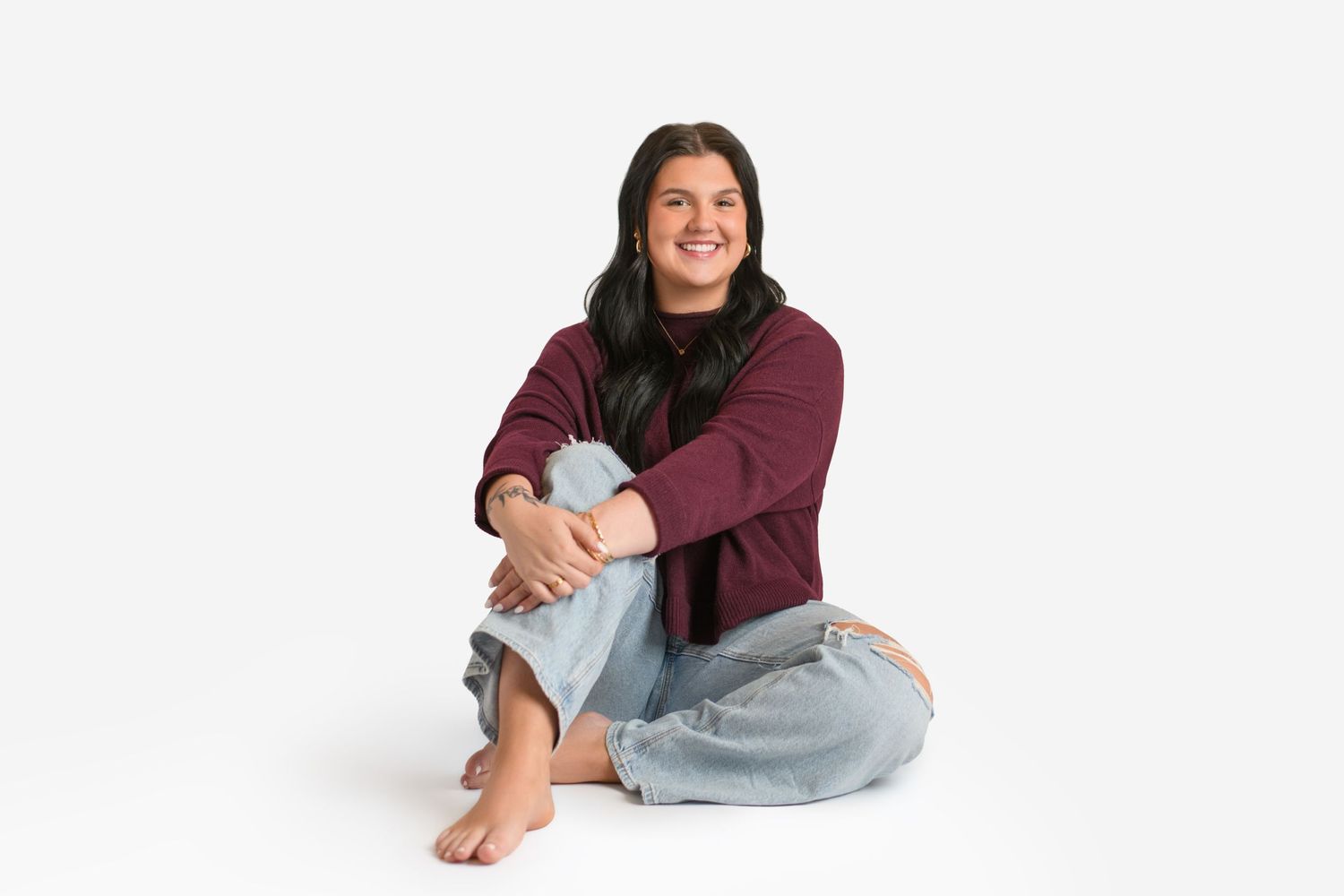 Person sitting cross-legged on white background wearing burgundy sweater and light denim jeans.