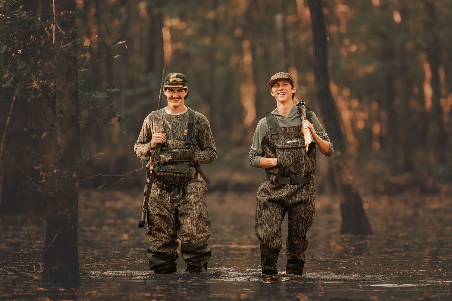 Two duck hunters in camouflage walk through a swamp area at sunset with rifles and tactical equipment.