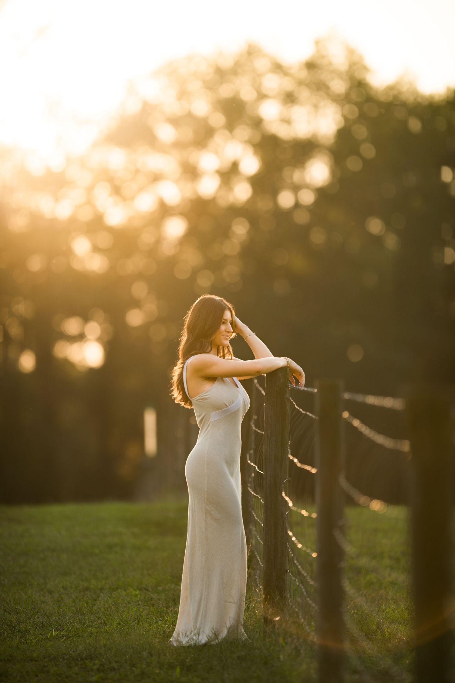 A woman in a long white dress stands by a wooden fence at sunset with dreamy golden bokeh lighting in the background.