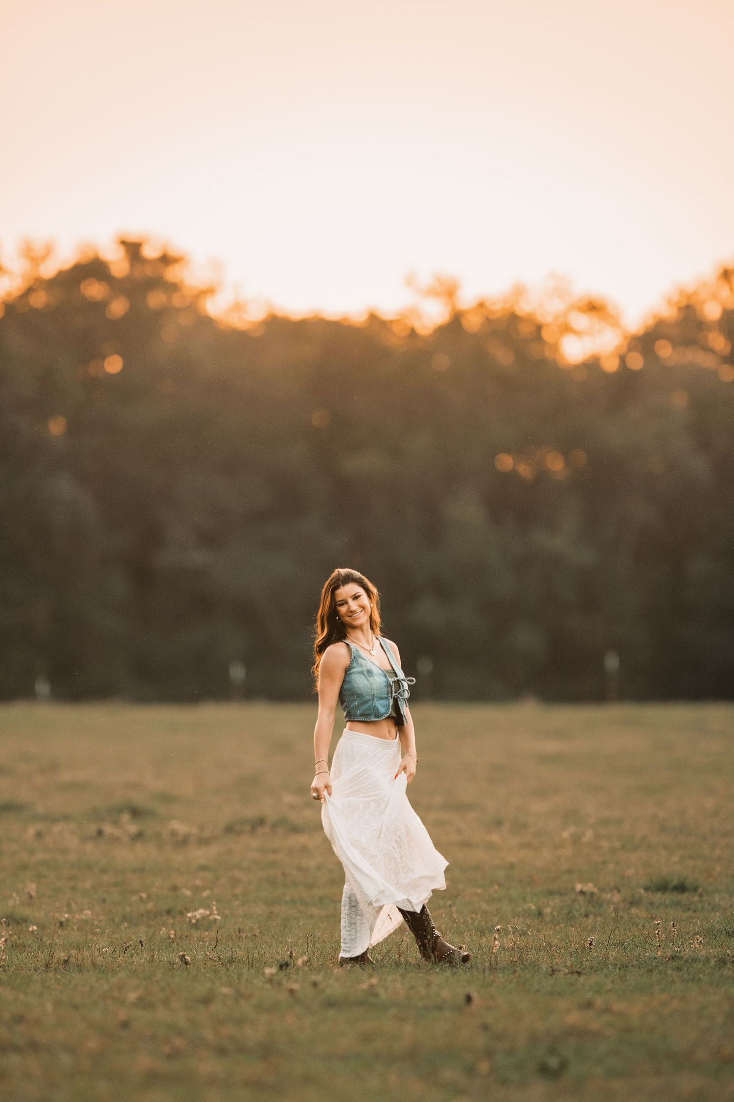 Young woman in flowy white skirt and denim top poses in golden field at sunset.