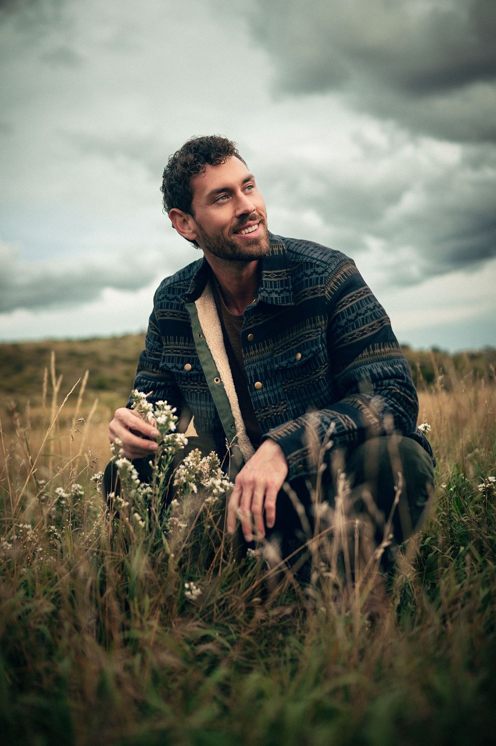Smiling man in patterned jacket crouching in golden grass field holding wildflowers under dramatic cloudy sky.