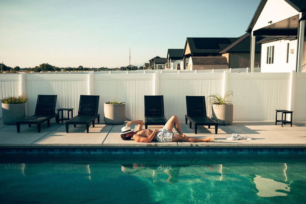 Person sunbathing poolside on a luxury outdoor patio with lounge chairs and white vinyl fence in a residential backyard.