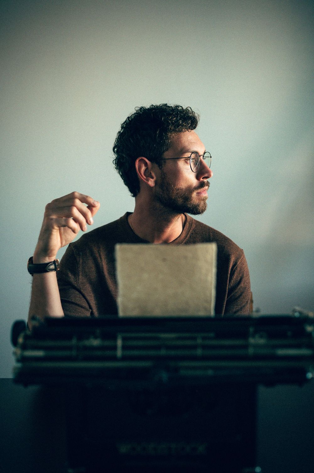 Man with curly hair and glasses sits pensively at a vintage typewriter, holding a pen in a moody studio setting.