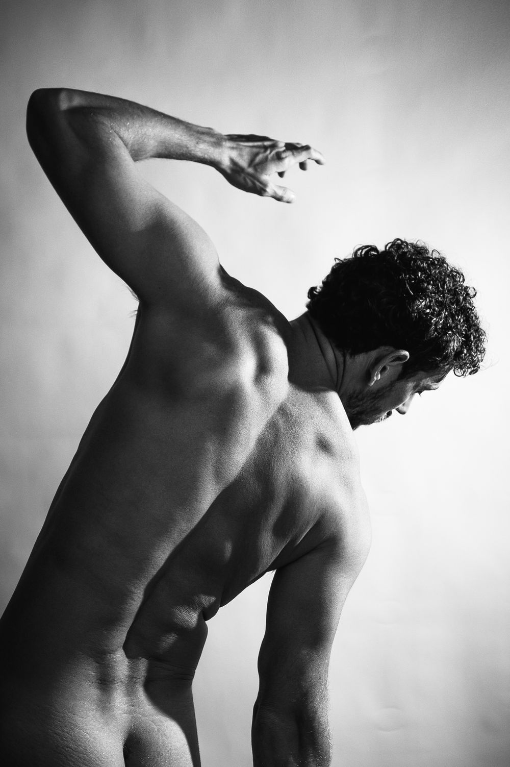 Black and white artistic photo of a muscular man posing with arm raised, showcasing back muscles in dramatic lighting.