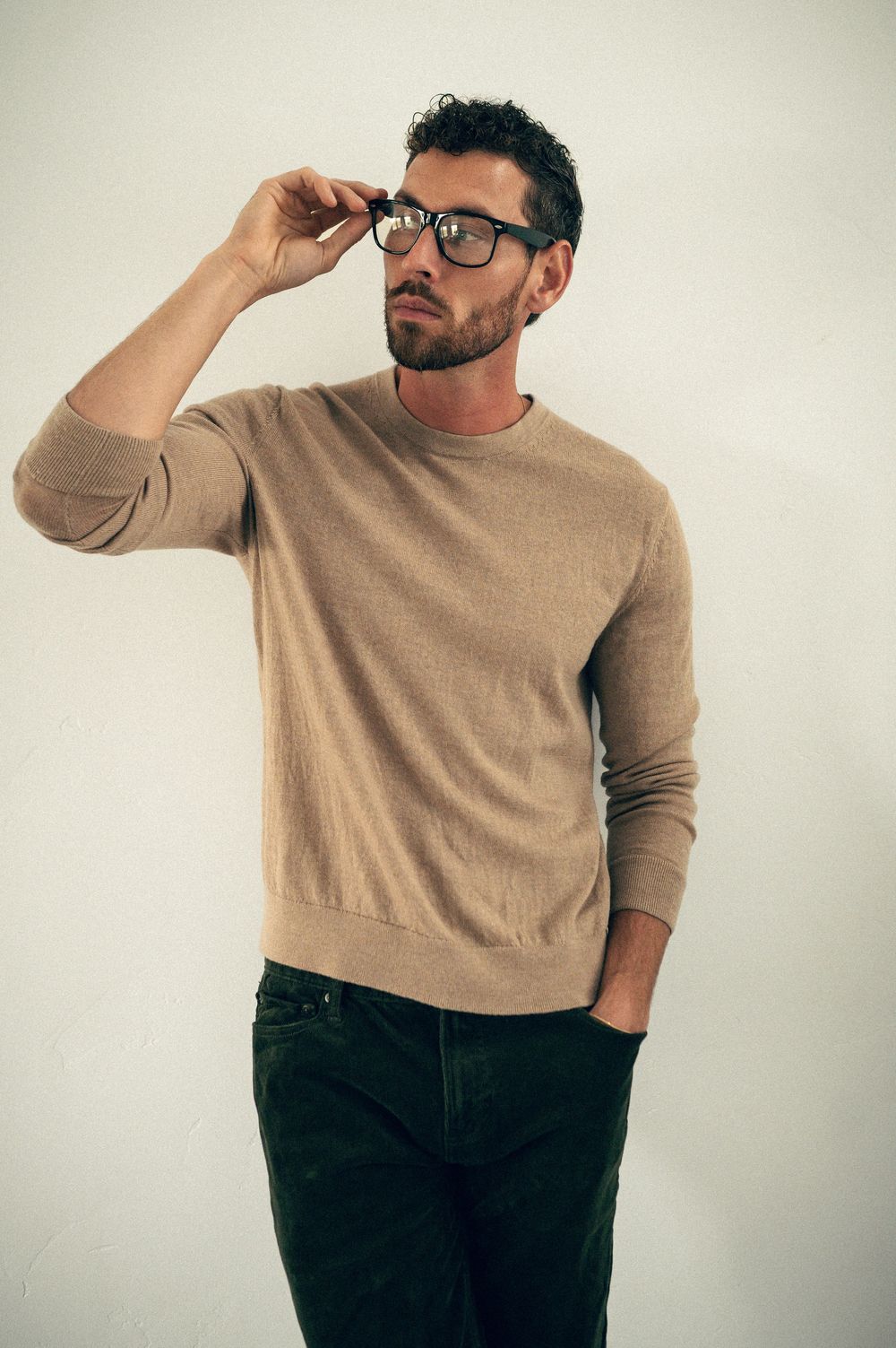 Stylish man in beige sweater and dark pants adjusting glasses against white background in casual fashion pose.
