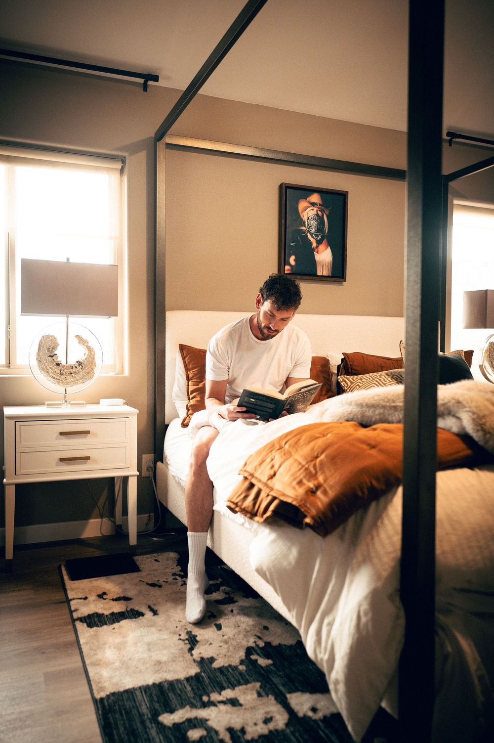 Man in white t-shirt sitting on a four-poster bed reading a tablet in a stylish, warmly lit bedroom.