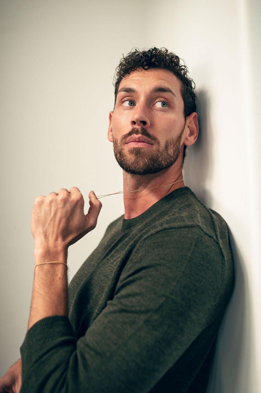 A thoughtful man with curly hair and beard in a green sweater leans against a wall, holding glasses contemplatively.