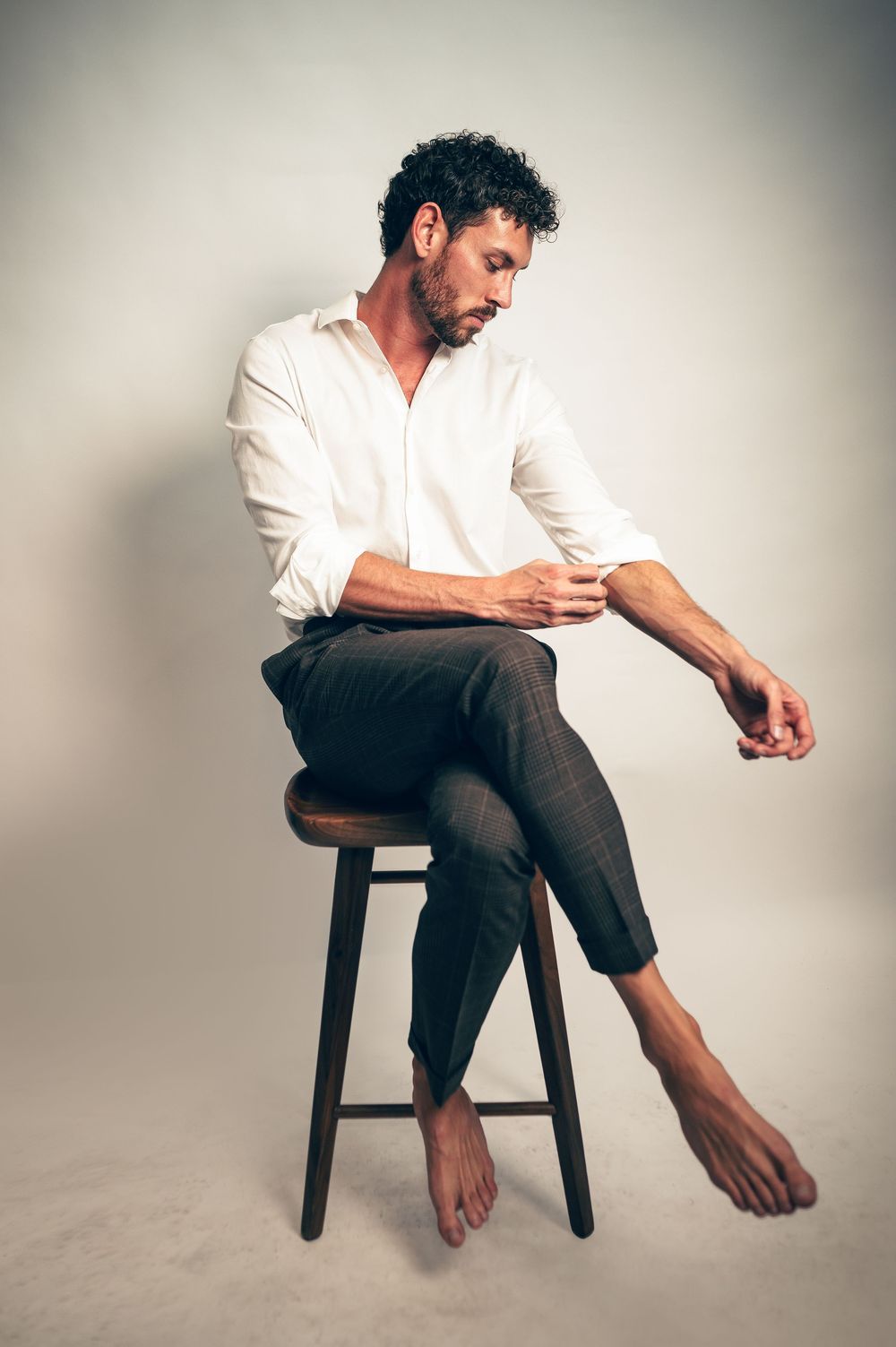 Barefoot man in white shirt and dark trousers sits on wooden stool, looking down in studio setting.