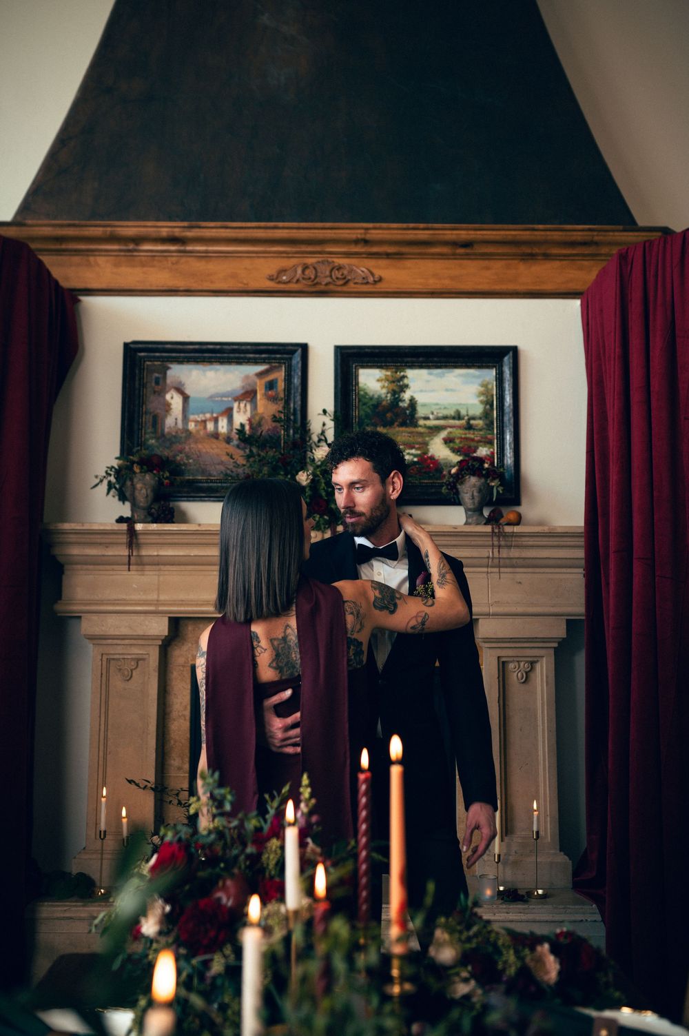 Couple sharing romantic moment by candlelit fireplace with Italian landscape paintings, man in suit with tattoos, woman in burgundy gown.