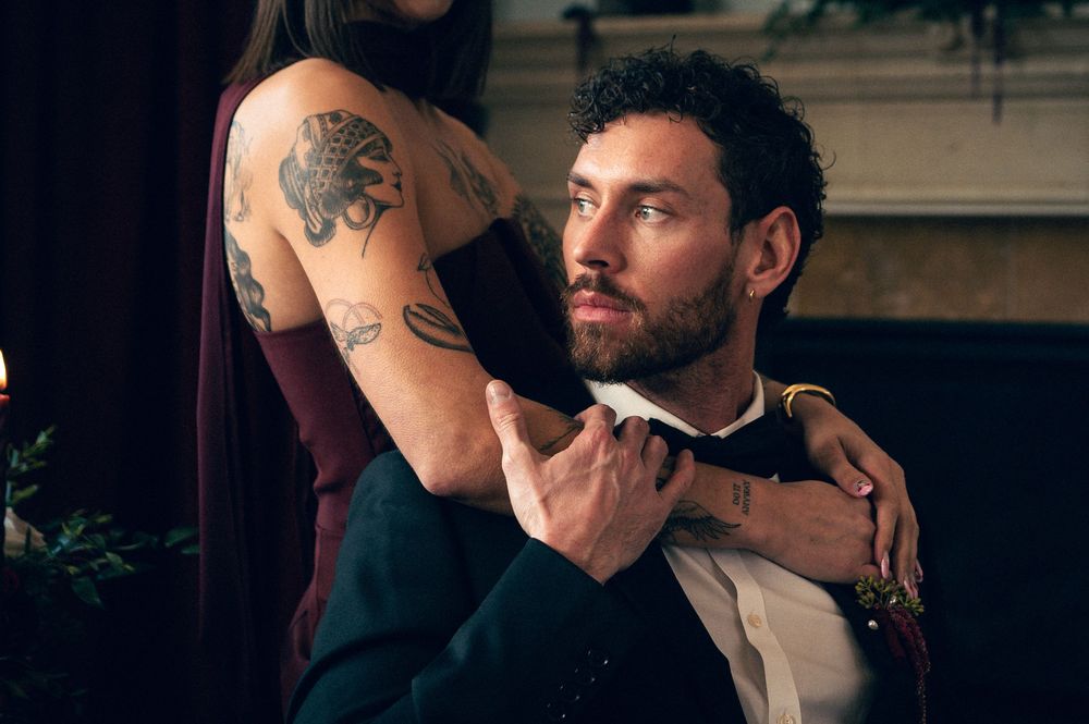 A bearded man in formal attire sits while a tattooed woman places her hands on his shoulders indoors.