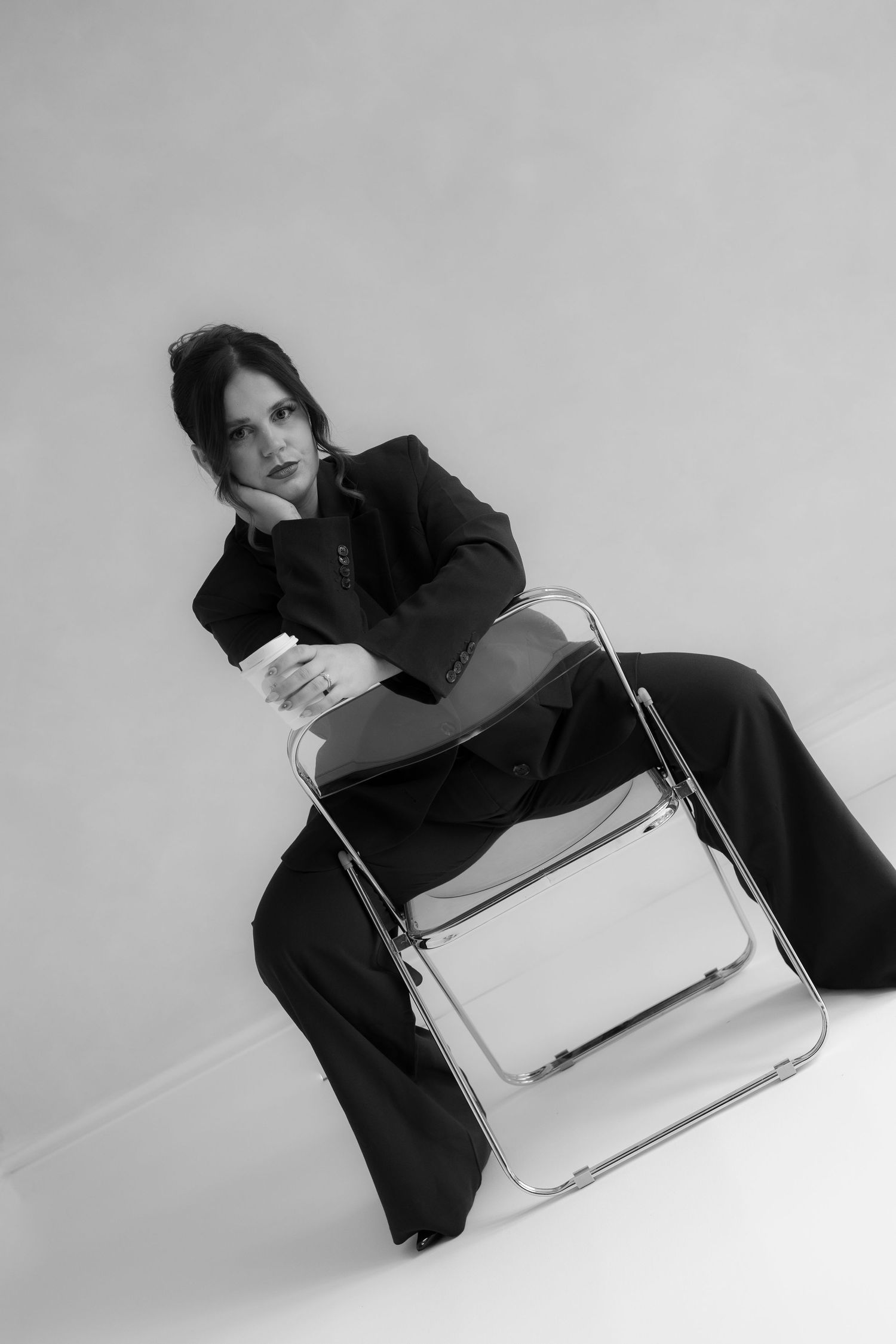 black and white image of photographer branding session while sitting backwards on a clear chair holding a coffee in studio setting.