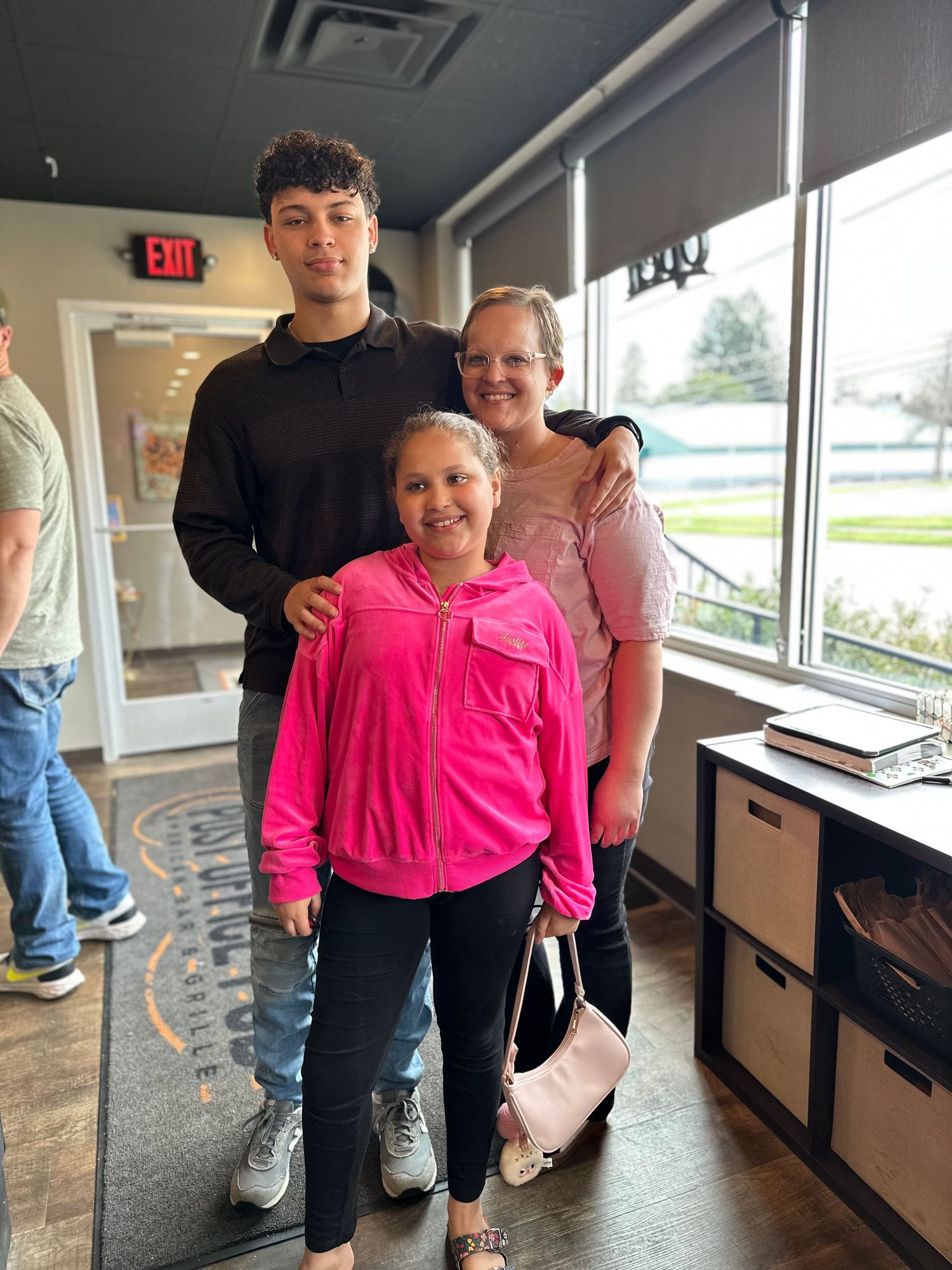 Three people posing together indoors, two wearing pink and one in black, smiling in a bright modern space.