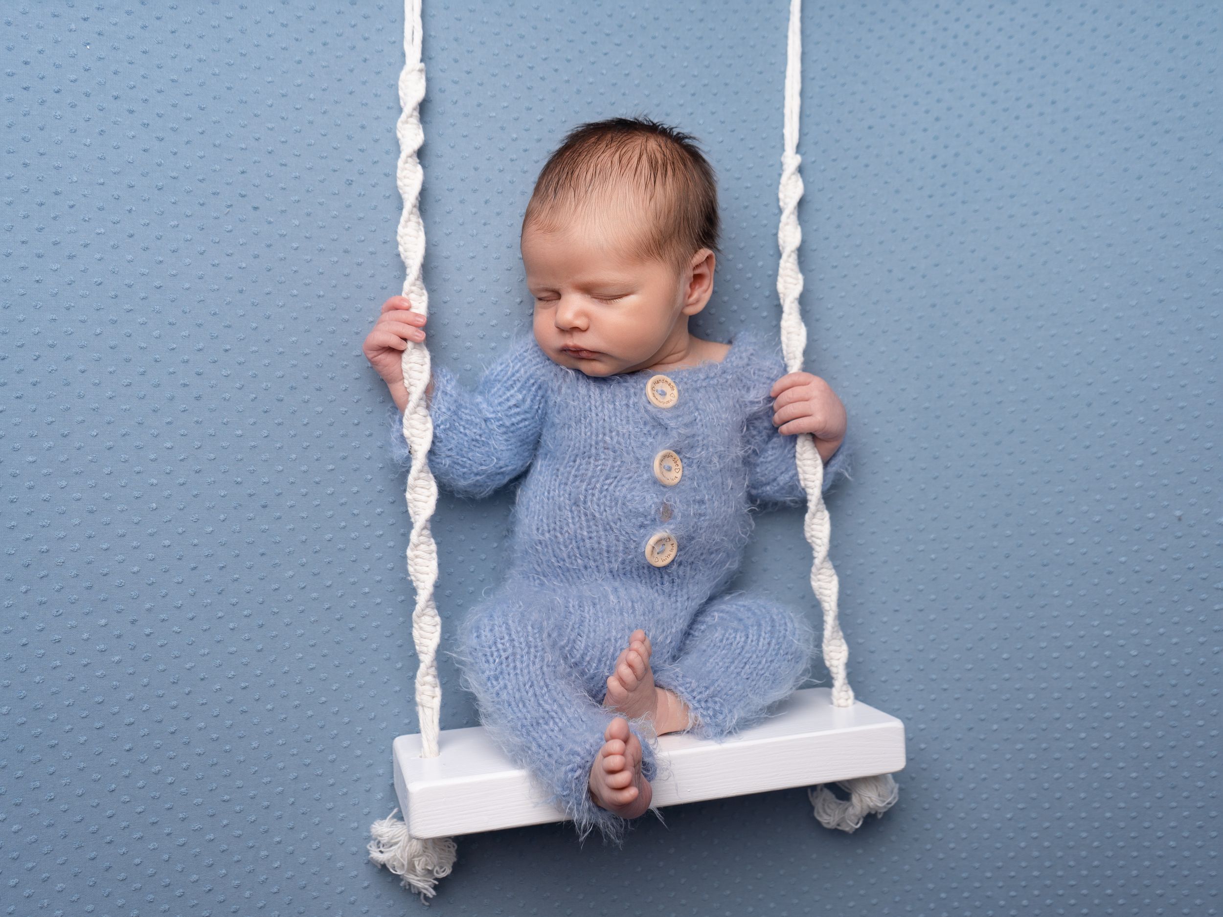 Newborn baby boy in blue fluffy romper on swing prop during newborn session with local photographer near Doncaster.