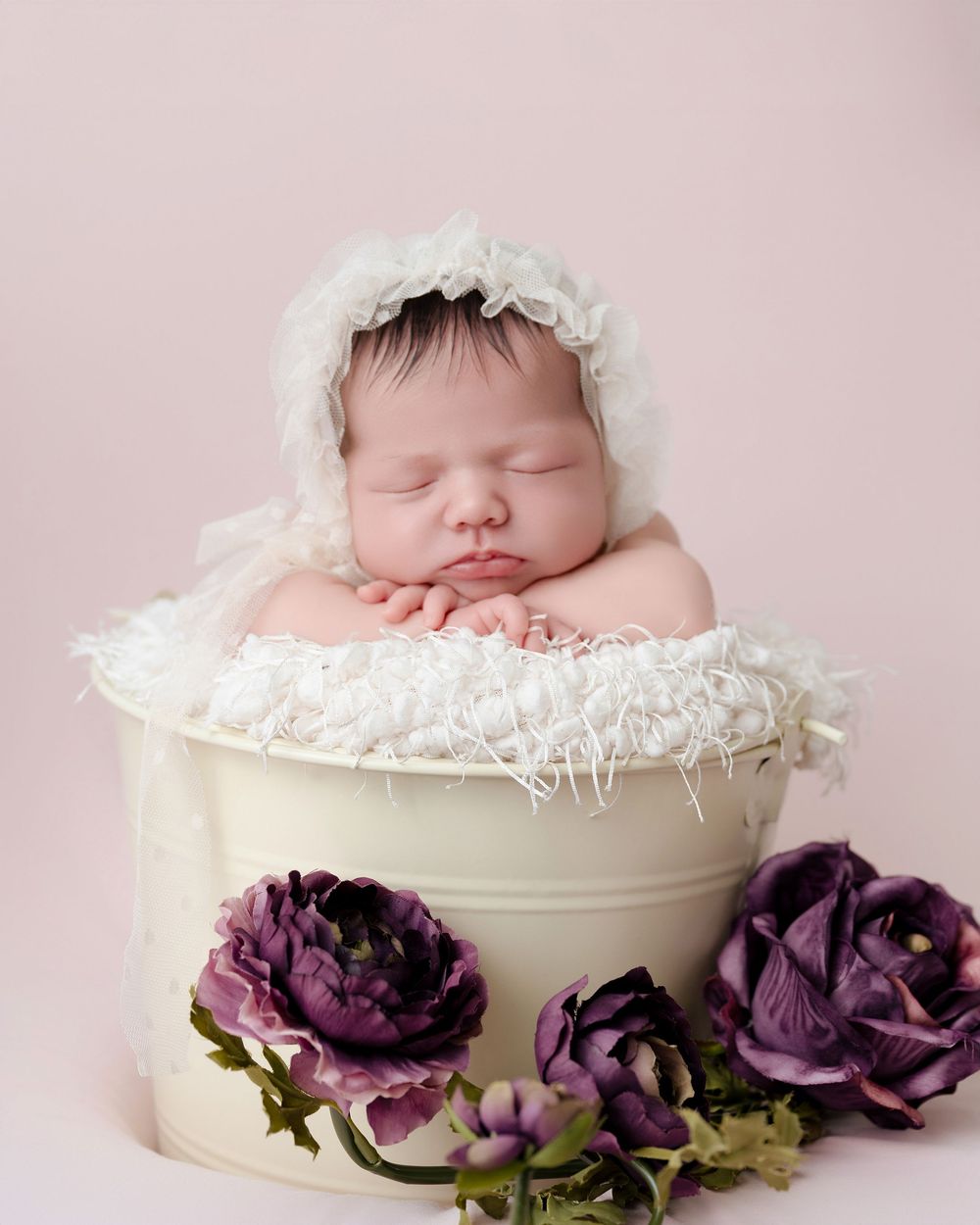 Sleeping newborn wearing white bonnet posed in cream container with purple flowers against pink background.