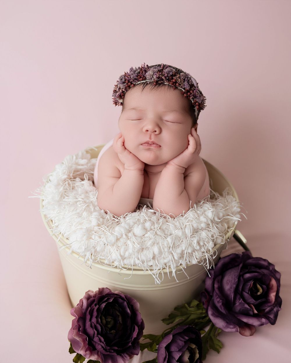 Sleeping baby with floral headpiece resting on cream container decorated with purple roses.
