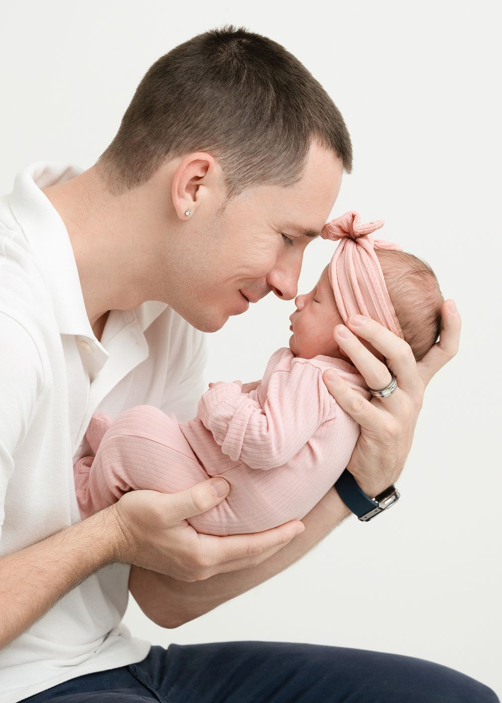 Parent in white shirt snuggles and kisses small baby in pink onesie against white background in tender moment.