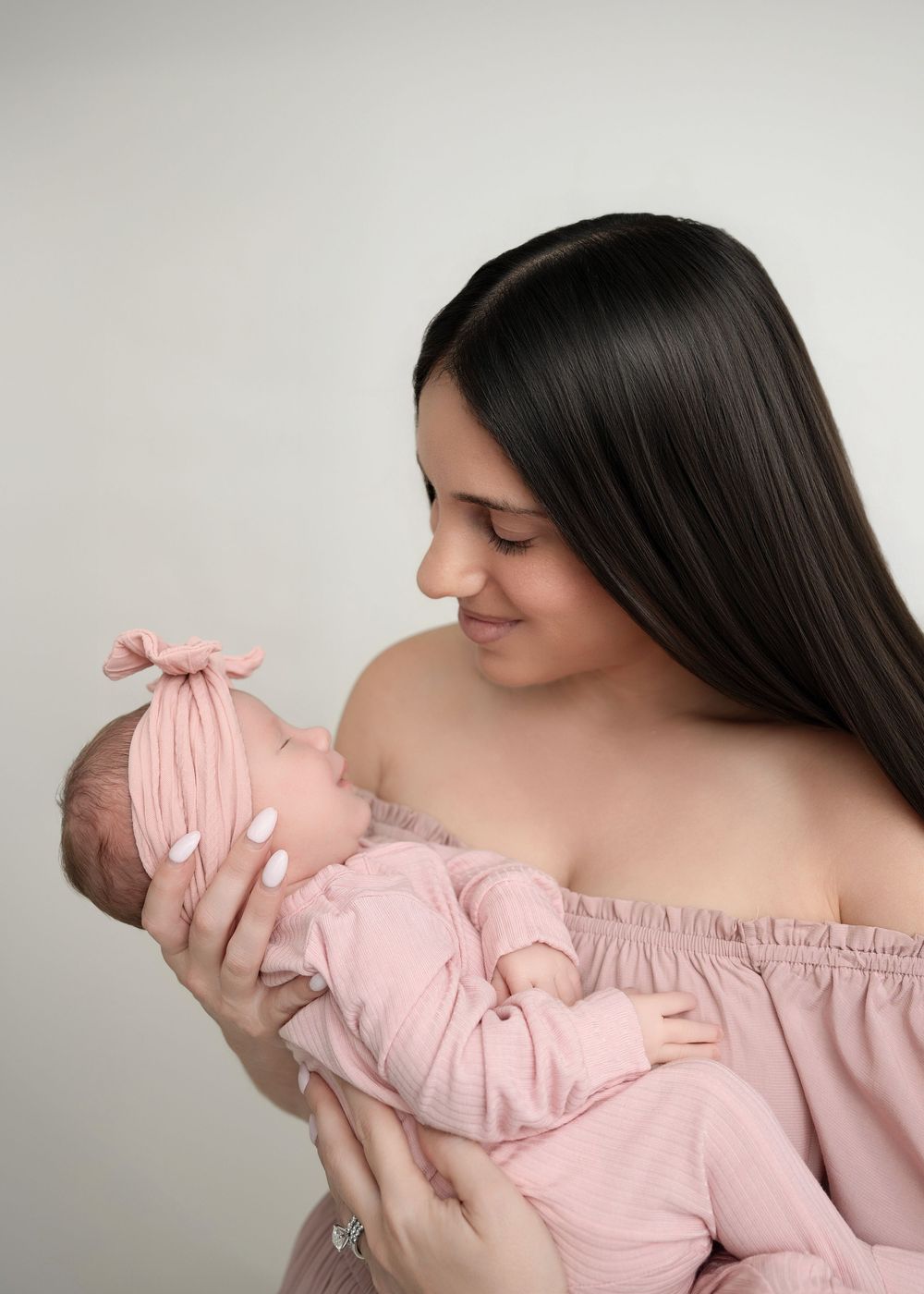 Woman in off-shoulder pink dress holds sleeping newborn baby in matching pink outfit in tender portrait series.