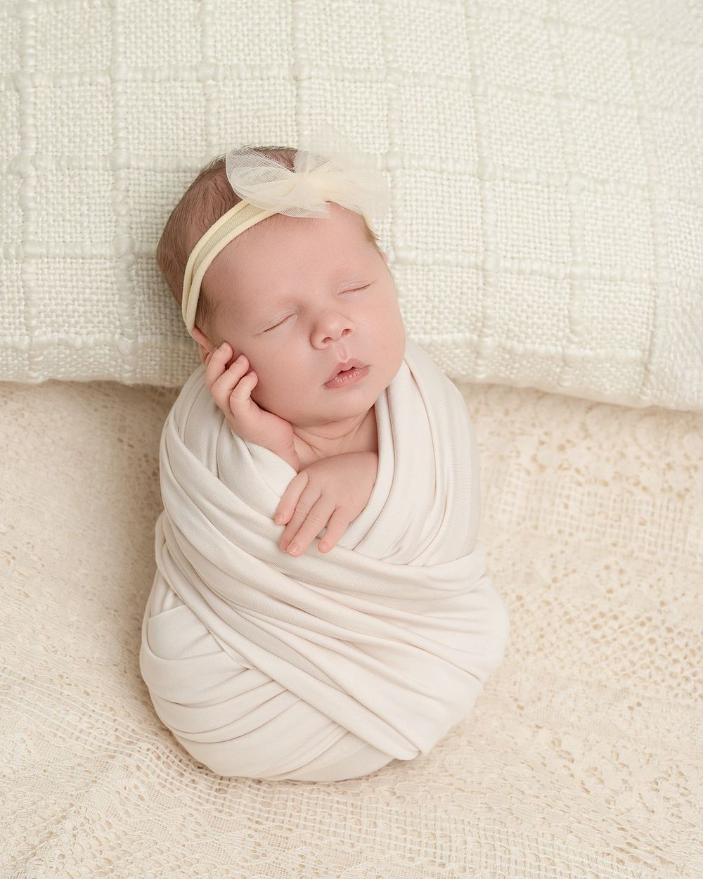 Sleeping newborn wrapped in soft white fabric against cream textured background in artistic portrait pose.