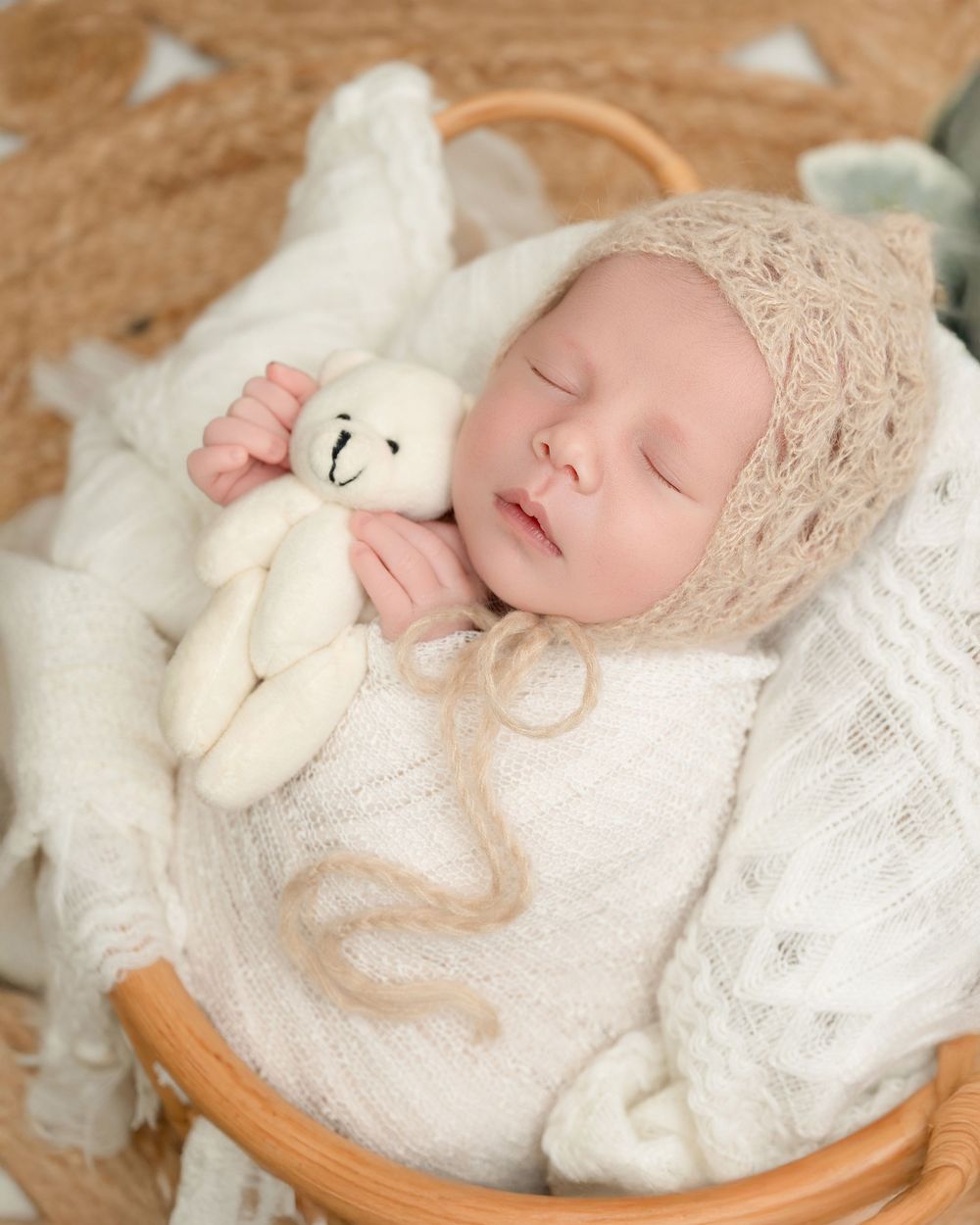 A sleeping newborn wrapped in white lace swaddle with plush toy in wooden bowl for professional photography session.