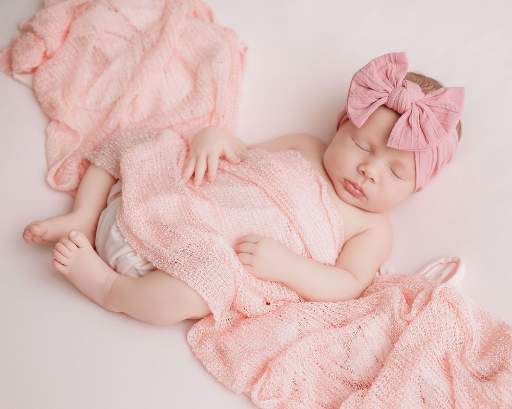 A sleeping newborn in pink lace outfit during professional photography session on light pink background.