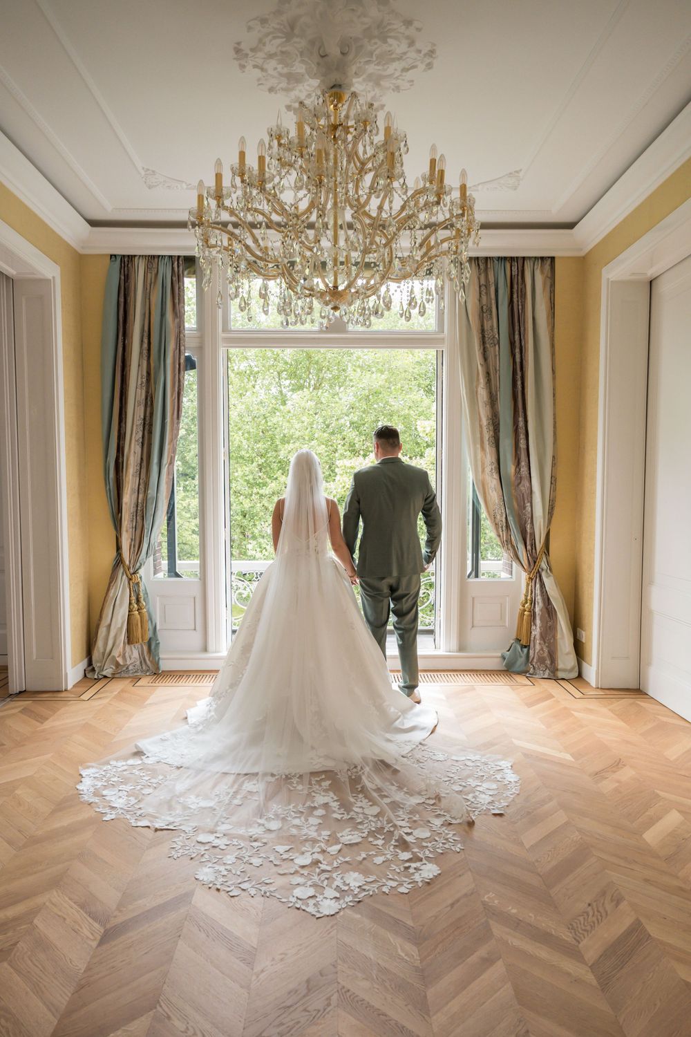 Bride and groom stand hand-in-hand before grand window in elegant ballroom with chandelier and herringbone floors.