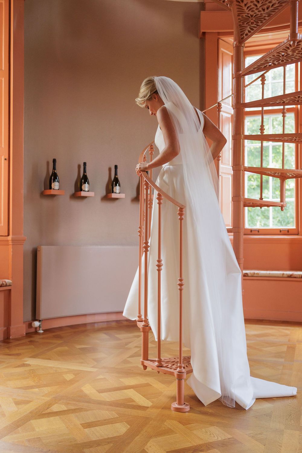 Bride in white gown and veil stands at wooden staircase railing in elegant salmon-toned room with wine bottles on wall.