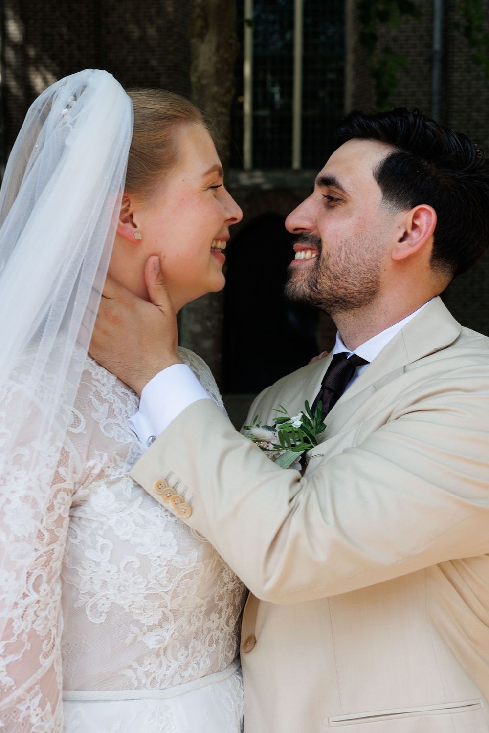 A smiling bride in lace dress and veil gazes at groom in beige suit with boutonniere at wedding ceremony.