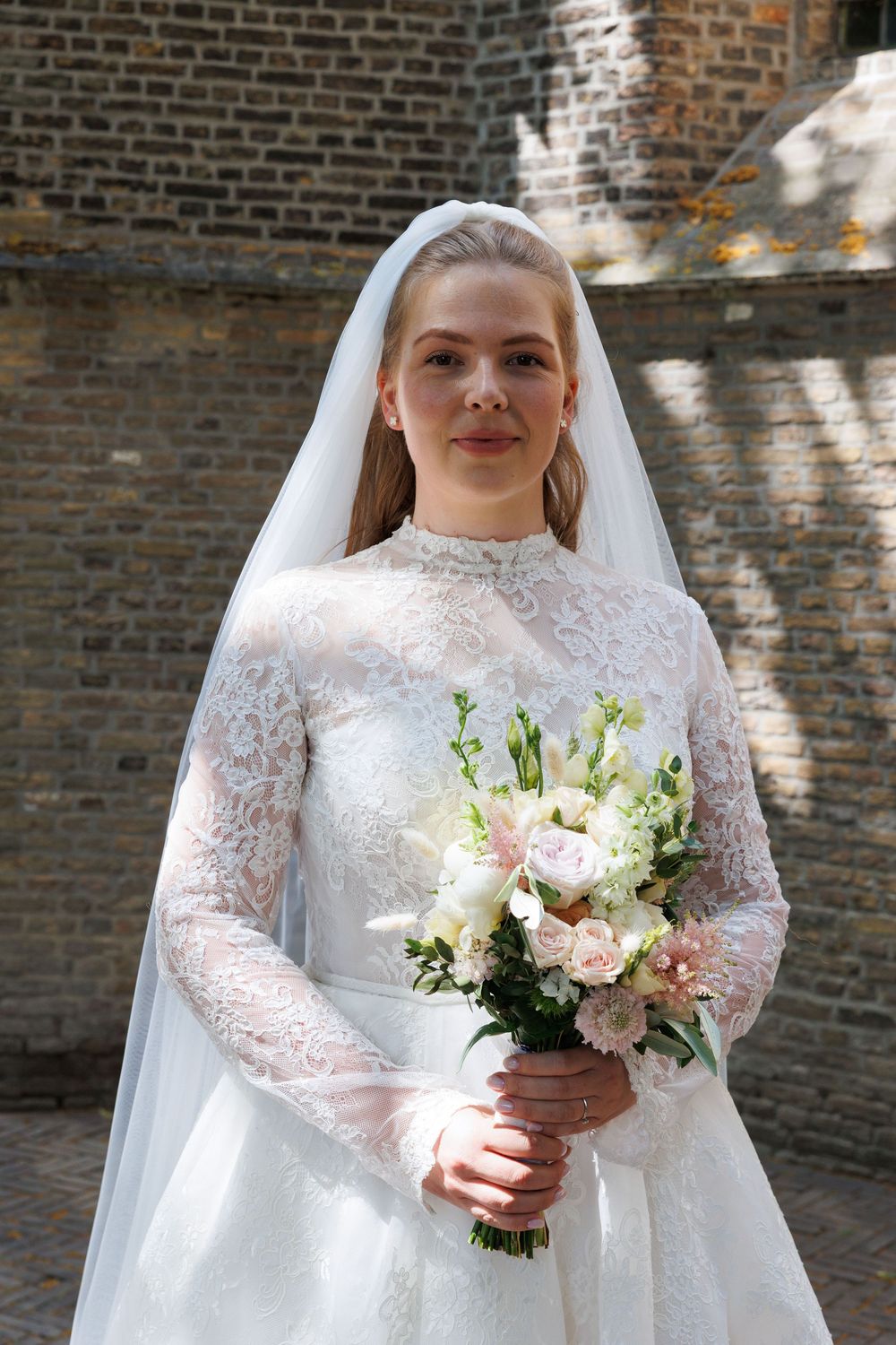Young bride in white lace gown and veil holding pink and white bouquet, standing before brick wall.