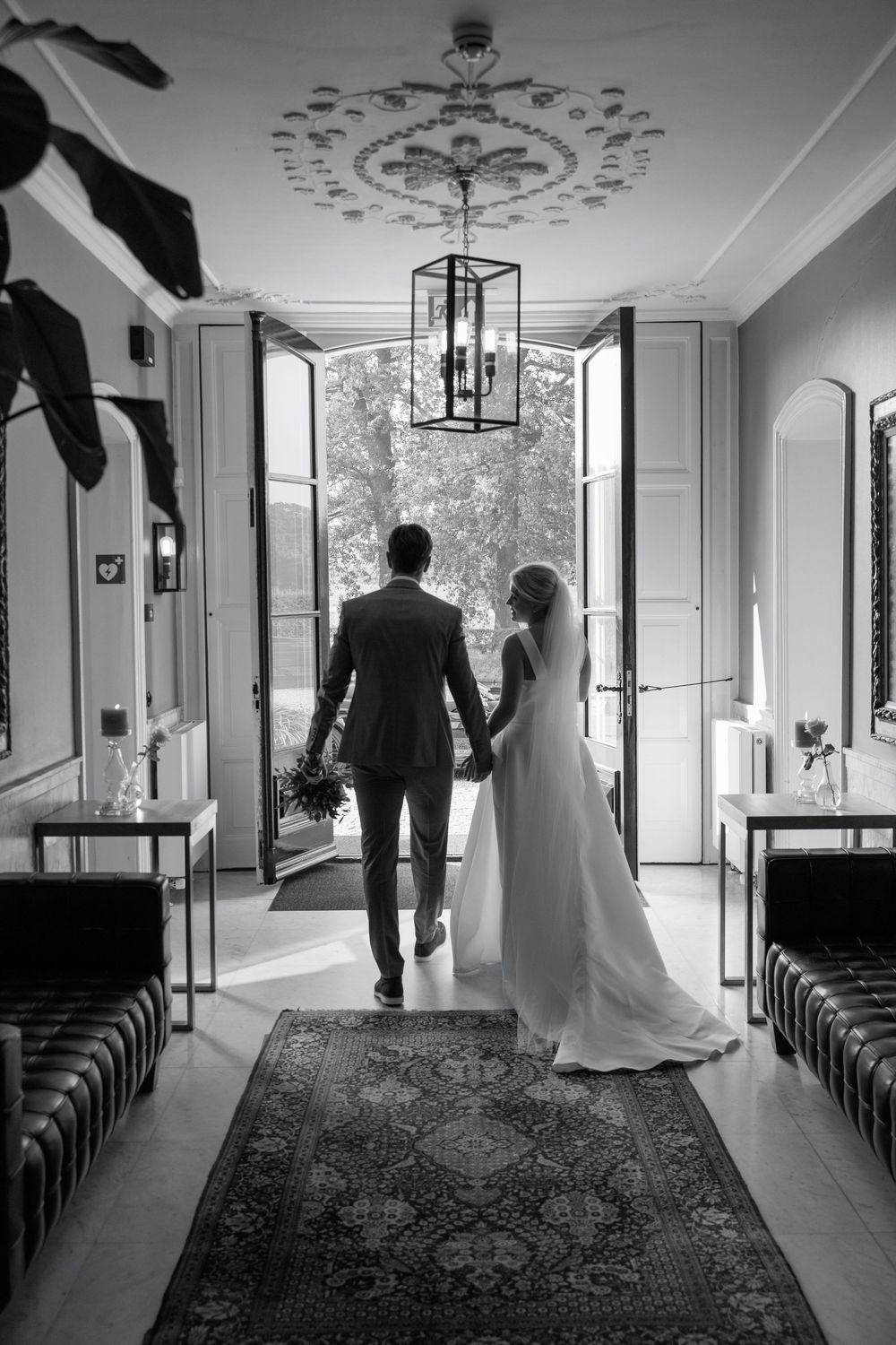 Black and white photo of bride and groom holding hands walking toward open doors at elegant wedding venue.