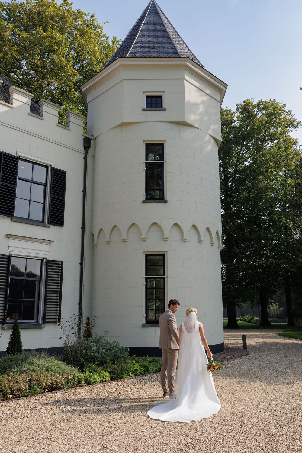 Bride and groom standing before a white castle tower on their wedding day, surrounded by lush greenery.