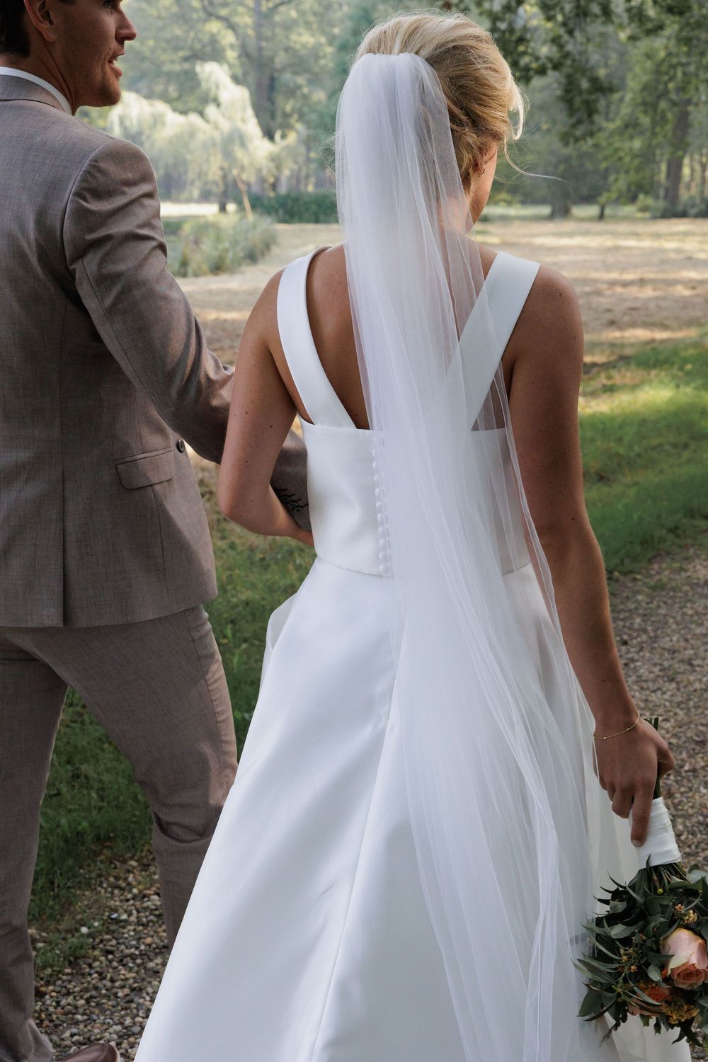 Bride in white gown with veil walks with groom in tan suit through scenic outdoor wedding venue.