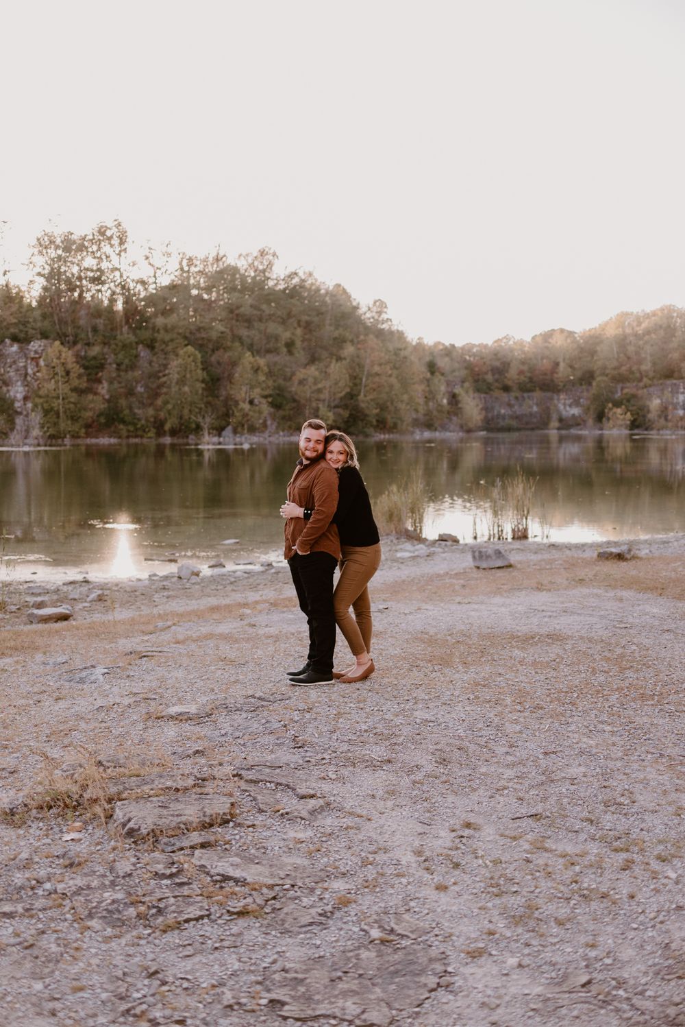 Wide landscape shot of a couple embracing on a rocky shoreline near an autumn lake.