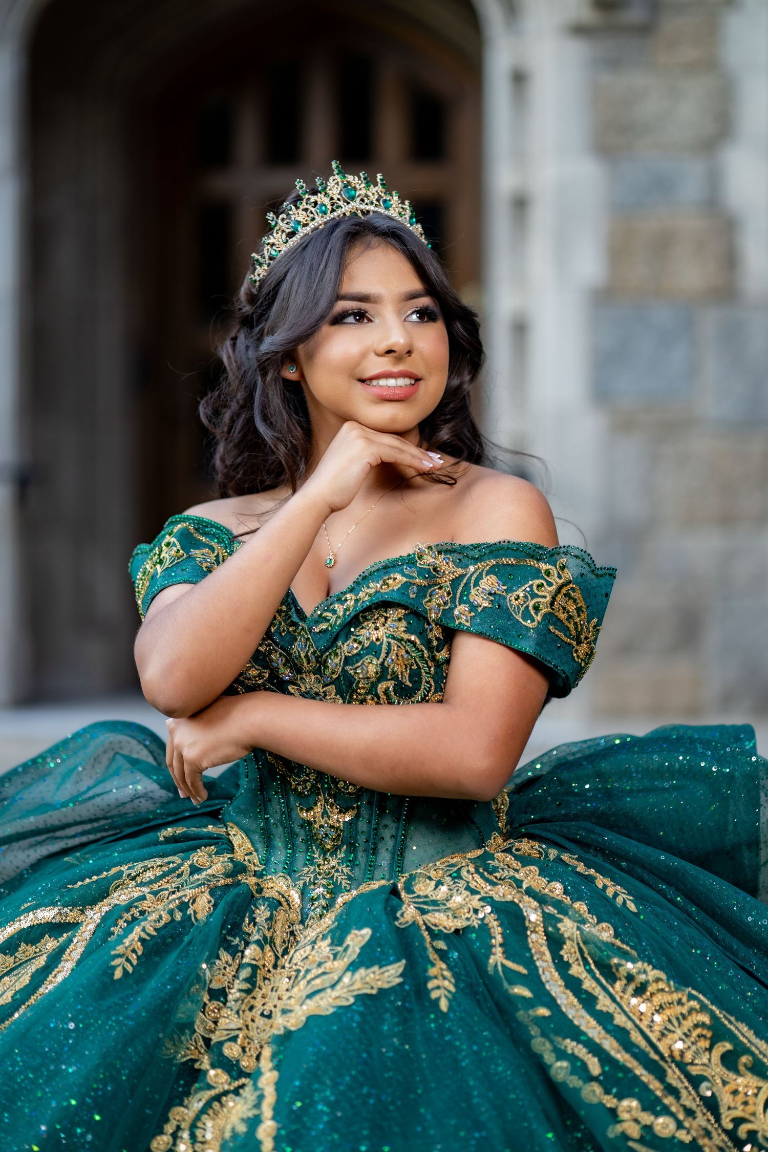 Young woman in an emerald green and gold embroidered quinceañera gown wearing a crown poses outdoors.