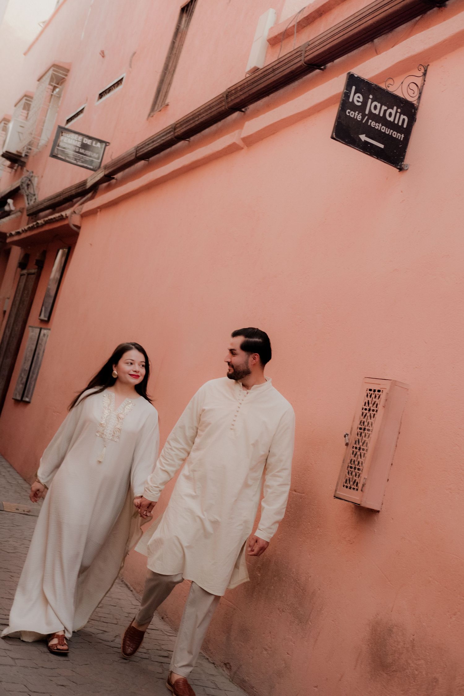 Couple in matching white traditional outfits holding hands beside a brick pillar.