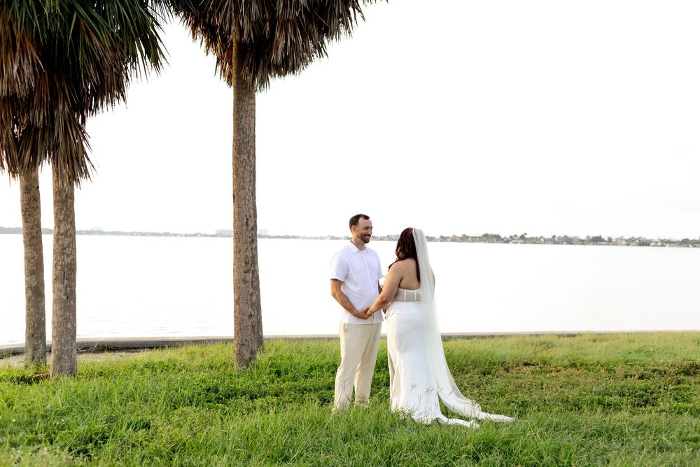 A couple in white stands by palm trees along a waterfront during their romantic outdoor wedding ceremony.