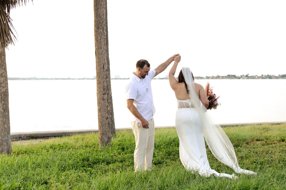 Dancing couple in white outfits share a romantic moment on grassy waterfront near palm trees at sunset.